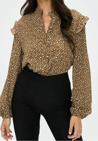 Blouse marron avec des taches, dotée d'un front à boutons, d'un col volanté et de manches longues. Portée avec un pantalon noir taille haute. Texture de tissu douce.
