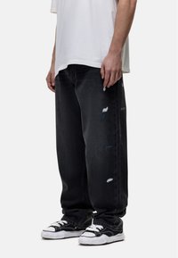 Jeans in denim nero con una vestibilità ampia, caratterizzati da dettagli strappati e sottili toppe in contrasto. Indossati con sneakers nere e una camicia bianca.