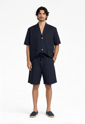 Uomo che indossa una camicia blu navy a maniche corte con bottoni e due tasche frontali, pantaloncini con coulisse abbinati e scarpe slip-on blu navy, in piedi su sfondo bianco.