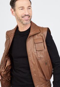 Braune, lederne ärmellose Weste mit einem Reißverschluss vorne, mehreren Taschen und geripptem Saum. Getragen über einem schwarzen Langarmshirt. Glatte Textur.