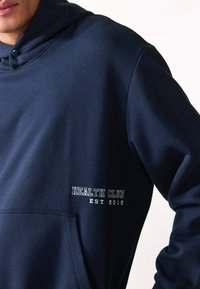 Navy blue hooded sweatshirt with a front pocket, featuring white "HEALTH CLUB" and "EST 2012" text in a simple font. Soft, cotton material.