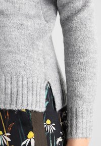 Grauer Strickpullover mit geripptem Saum und seitlichen Schlitzen. Er hat eine weiche Textur und wird mit floral gemustertem Stoff kombiniert.