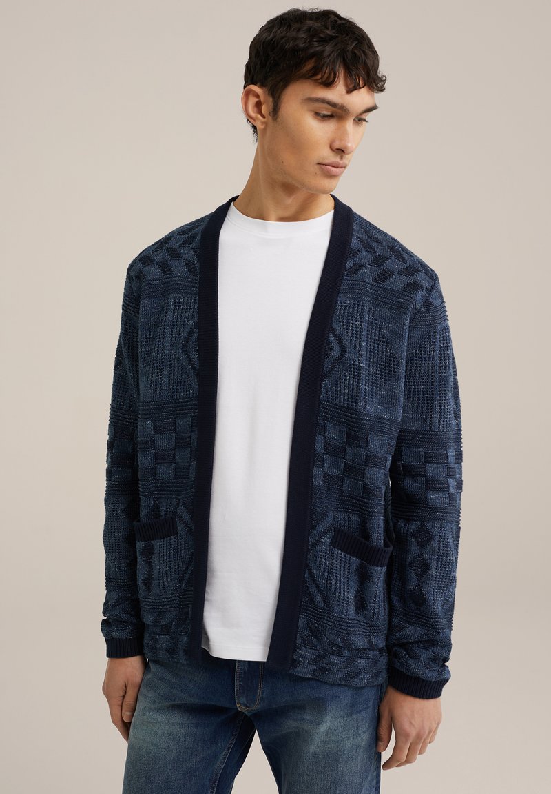 Marineblauwe gebreide cardigan met textuurpatronen, contrasterende donkere afwerking en voorzakken, gedragen over een witte t-shirt.