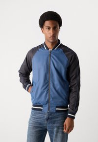 Blaue und schwarze Bomberjacke mit Reißverschluss, Rippbündchen und -saum, mit seitlichen Taschen und kontrastierenden Streifen entlang der Ärmel. Glatte Textur.