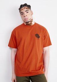 Camiseta de algodón color óxido con cuello redondo, mangas cortas y un gráfico negro en la parte izquierda del pecho. Textura suave.