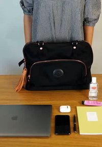 Black fabric laptop bag with rose gold accents, zipper, and tassel; features front pocket with logo; placed on a wooden surface beside tech items.