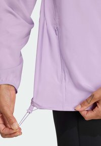 Veste légère lilas avec une poche latérale zippée et un ourlet ajustable par cordon. Texture lisse et poignets élastiques.