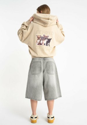 Kobieta w beżowej bluzie z kapturem z czterema małymi ilustracjami psów i napisem „Beyond Cuteness”, zestawiona z szarymi szerokimi szortami dżinsowymi.