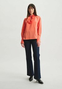 Chemise corail boutonnée avec détail de col à volants, associée à un jean évasé sombre et des escarpins noirs à bouts pointus, sur un fond neutre.