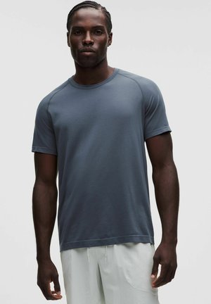 Uomo con capelli intrecciati indossa una camicia grigio scuro a maniche corte e pantaloni chiari su uno sfondo chiaro e uniforme.