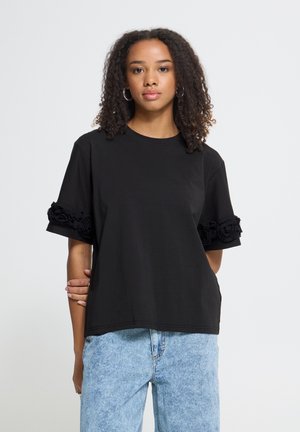 Femme debout portant un t-shirt noir à manches courtes avec des détails floraux sur les manches et un jean bleu clair, fond neutre, de face.