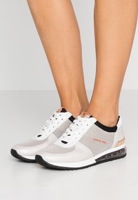 Białe i srebrne sportowe sneakersy z siateczkowymi panelami, okrągłym noskiem, czarnymi akcentami i przezroczystym obcasem. Posiadają system sznurowania oraz metaliczne detale.