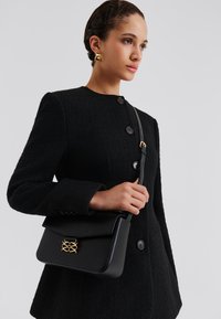 Μαύρη δερμάτινη γυναικεία τσάντα με διακοσμητικό χρυσού χρώματος και κλείσιμο με καπάκι. Διαθέτει δομημένο σχέδιο και μακριά, ρυθμιζόμενη τιράντα.