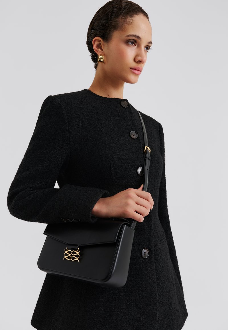 Μαύρη δερμάτινη γυναικεία τσάντα με διακοσμητικό χρυσού χρώματος και κλείσιμο με καπάκι. Διαθέτει δομημένο σχέδιο και μακριά, ρυθμιζόμενη τιράντα.