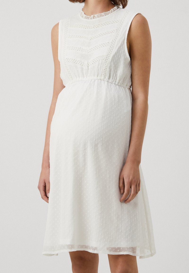 Robe blanche sans manches longueur genou en tissu texturé, détails en dentelle sur le corsage et taille froncée, portée par une personne debout.