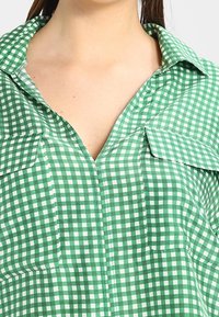 Chemise à carreaux gingham vert et blanc avec un col, deux poches avant et une patte de boutonnage. Tissu léger avec une texture lisse.