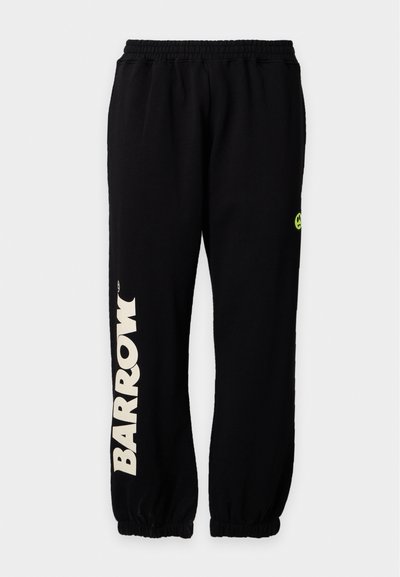 Pantalon de survêtement noir avec taille et poignets élastiques, "BARROW" imprimé verticalement en grandes lettres blanches sur la jambe gauche, et un petit visage souriant jaune sur la jambe droite.