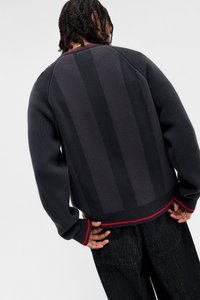 Mörkgrå stickad tröja med texturerade vertikala ränder, ribbade muddar och röda detaljer vid nederkanten. Oversized passform med raglanärmar.