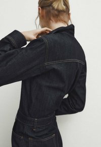 Blondynka widziana z tyłu, poprawiająca kołnierz ciemnej dżinsowej kurtki z widocznymi szwami i tylnymi kieszeniami.