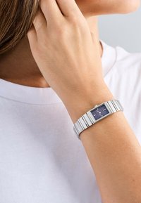 Orologio da polso rettangolare in metallo argento con quadrante blu scuro, caratterizzato da indici ore argentati e un bracciale in metallo flessibile. Sfondo di una maglietta bianca.