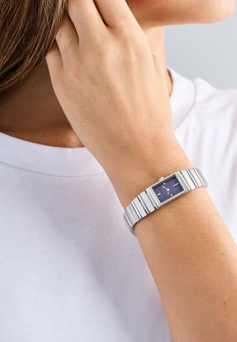 Orologio da polso rettangolare in metallo argento con quadrante blu scuro, caratterizzato da indici ore argentati e un bracciale in metallo flessibile. Sfondo di una maglietta bianca.