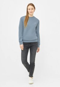 Lichtblauwe sweatshirt met een ronde hals en ribbels aan de mouwen, gecombineerd met donkergrijze slim fit jeans en witte sneakers, gedragen door een model.