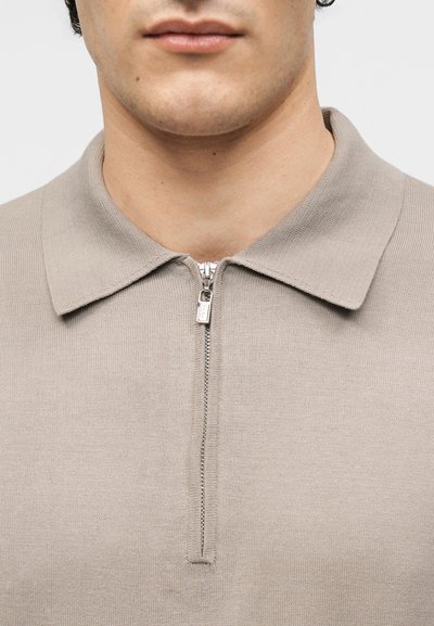 Jeune adulte portant un polo en maille beige avec une fermeture éclair argentée partiellement fermée au niveau du cou.
