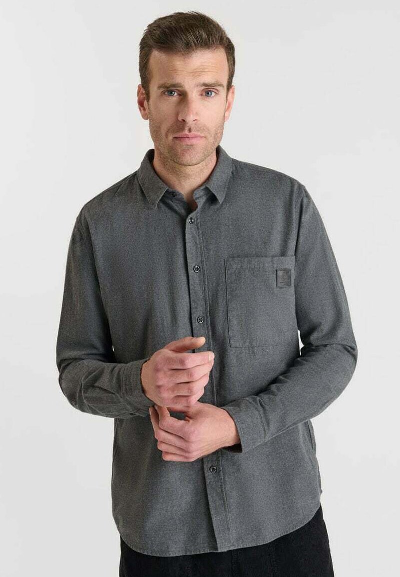 Camisa de botón gris oscuro en tejido texturizado, con un bolsillo que lleva un parche de logo y un cuello, mangas largas abotonadas de manera ordenada en las muñecas.