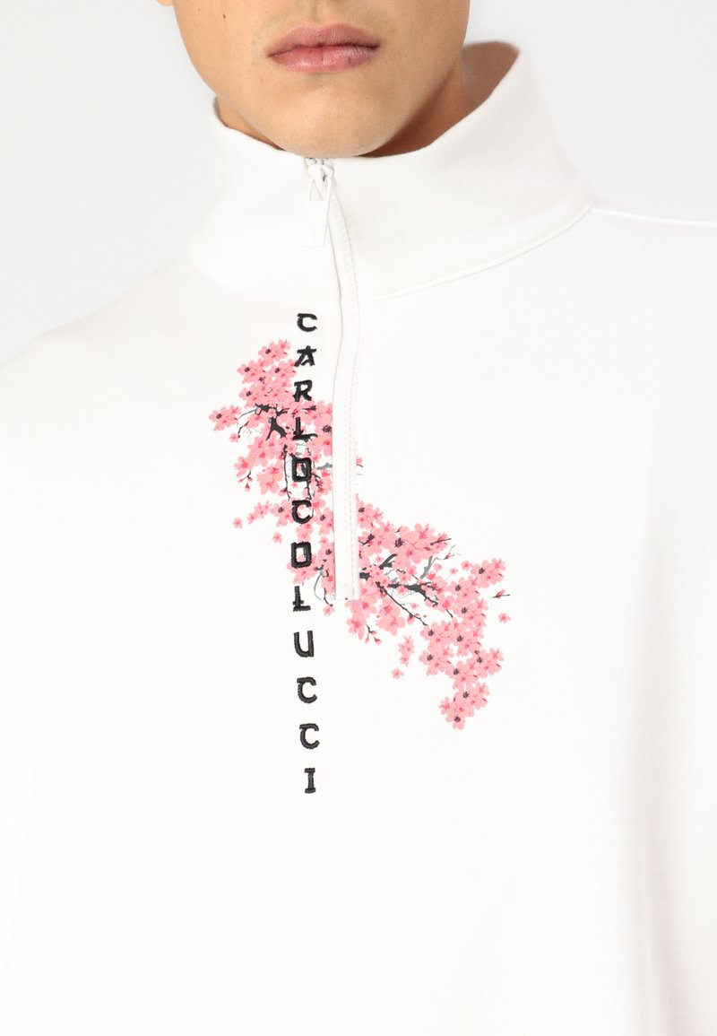 Biała bluza z zamkiem błyskawicznym z różową haftowaną florystyką i napisem "CARLO COLUCCI" pionowo po lewej stronie. Miękki, casualowy design.