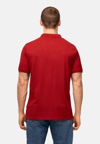 Polo shirt in cotone rosso con colletto e maniche corte, caratterizzato da una texture liscia e un taglio dritto, indossato con jeans in denim blu.