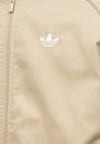 Veste beige avec une fermeture éclair centrale et un logo Adidas Trefoil blanc brodé sur la poitrine.