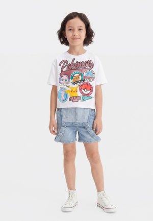 Enfant portant un t-shirt blanc avec un motif Pokémon avec des personnages et un short en denim avec des bretelles, debout devant un fond uni.