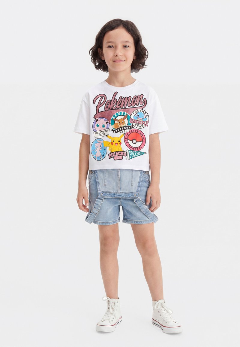 Παιδί που φοράει λευκό T-shirt με γραφικά Pokémon με χαρακτήρες και τζιν σορτς με τιράντες, στέκεται μπροστά σε απλό φόντο.