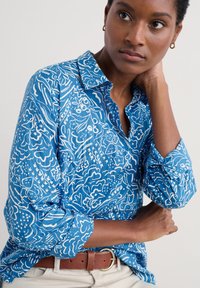 Blå skjorta med knappar och ett vitt blommönster. Tillverkad av lättviktsmaterial med uppvikta ärmar. Brun bälte som accent.