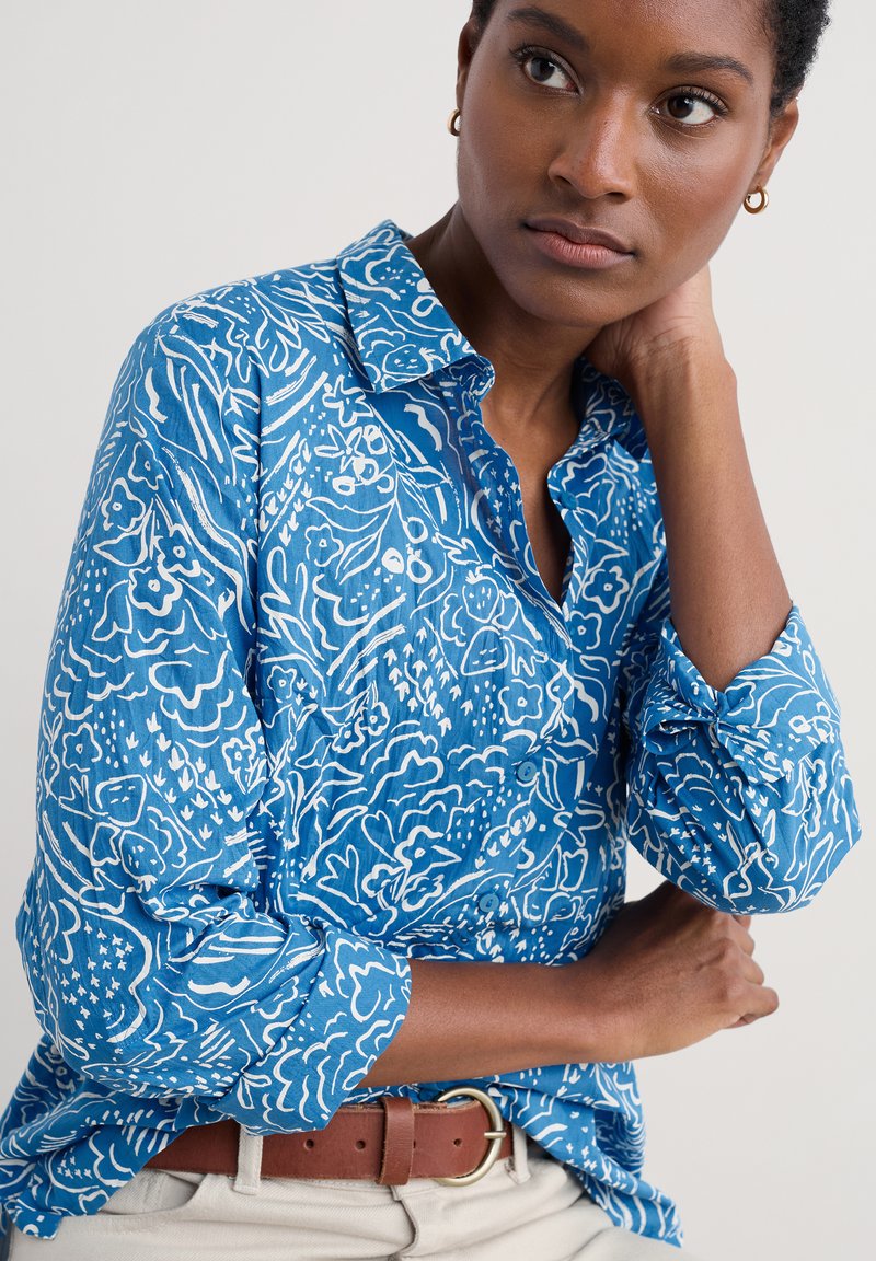 Blå skjorta med knappar och ett vitt blommönster. Tillverkad av lättviktsmaterial med uppvikta ärmar. Brun bälte som accent.