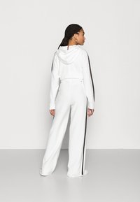 White hoodie and matching wide-leg pants, featuring brown side stripes, soft texture, and a drawstring hood. Back view highlights relaxed fit.
