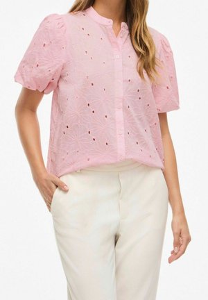 Woman wearing a light pink button-up blouse with puff sleeves and eyelet floral embroidery, paired with high-waisted white pants.