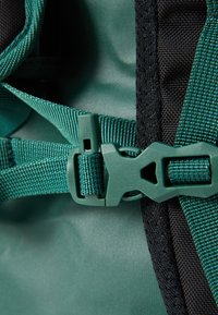 Green and black fabric backpack close-up featuring intricate webbing, plastic buckle, textured surface, and contrasting design elements.