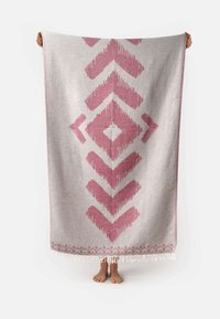 Gran tejido de color gris claro con un patrón geométrico en rosa, que presenta flechas y diamantes, y bordes con flecos, sostenido por una persona.