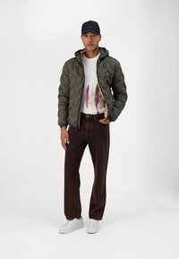 Giacca trapuntata verde oliva con cappuccio, maglione bianco con macchie marroni, pantaloni marroni e sneakers bianche. Design semplice e colori neutri.