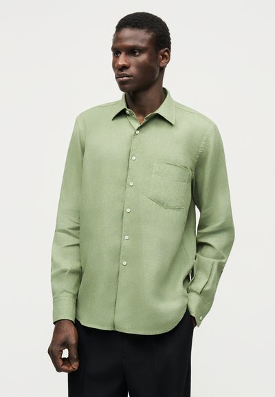 Άνδρας που φοράει ένα ανοιχτοπράσινο μακρυμάνικο πουκάμισο με τσέπη στο στήθος, στέκεται με το ένα χέρι στην τσέπη μαύρου παντελονιού.