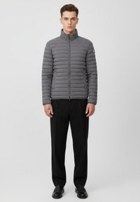 Grå puffjacka med quiltat mönster, hög krage, frampartizip och två sidofickor, i kombination med svarta byxor och skor.