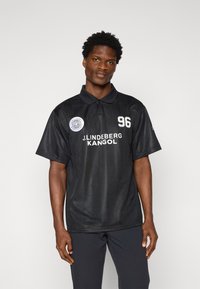 Sort polo-shirt lavet af letvægtsstof, med krave, lodrette striber, et cirkulært logo, teksten "J.Lindberg KANGOL" og tallet 96.