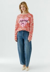 Junge Frau trägt einen rot-weiß gemusterten Pullover mit pinkem Herz und Schriftzug, kombiniert mit weitem blauen Jeans und weißen spitzen Schuhen.