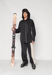 Skibekleidung mit einer schwarzen Kapuzenjacke mit Reißverschlusstaschen, schwarzen und beigen Hosen sowie einem senkrecht gehaltenen Snowboard mit Bindungen.