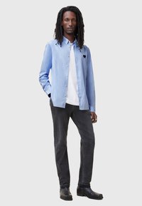 Hellblaues Hemd mit Knopfleiste und schwarzem Herzlogo, über einem weißen T-Shirt getragen, kombiniert mit dunkelgrauen Jeans und schwarzen Stiefeletten.