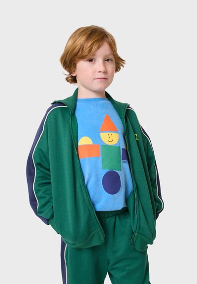 Zelený športový teplákový súbor s bielym lemovaním, doplnený modrým tričkom s farebnými geometrickými tvarmi: oranžový trojuholník, zelený štvorec, modrý kruh.