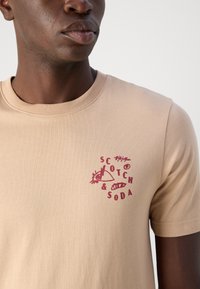Beige Baumwoll-T-Shirt mit Rundhalsausschnitt, das einen roten Grafikdruck zeigt. Das Design umfasst abstrakte Formen und den Text "SCOTCH & SODA."
