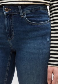 Σκούρα μπλε τζιν με κλασική γραμμή, διαθέτουν εμπρόσθια τσέπη, χάλκινο κουμπί και διακριτική φθορά στο μηρό.