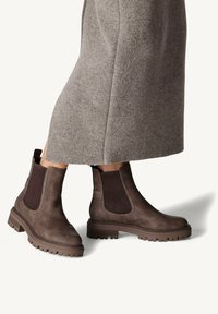 Stivali ankle in pelle marrone con pannelli elasticizzati laterali, suola in gomma robusta e finitura testurizzata; abbinati a un lungo capo grigio chiaro.